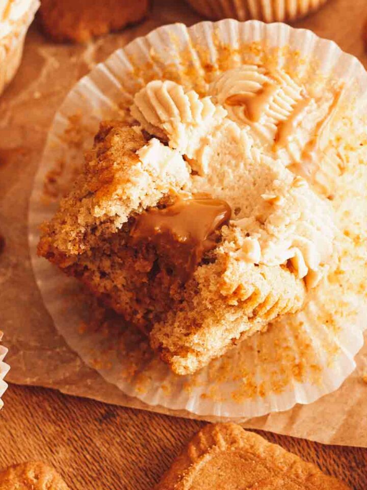 A closeup of a bitten Lotus Biscoff cupcake with Biscoff cookie butter filling dripping in the center on a wooden surface next to cupcakes and cookies.