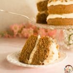 A close-up of a slice of carrot cake on a white plate with a two-layer carrot cake in the background.