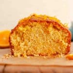 A close up of a cut orange loaf cake with orange zest on top on brown parchment paper.