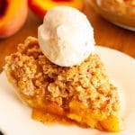 A slice of crumble top peach pie on a white plate topped with a ball of vanilla ice cream with peaches in the background.