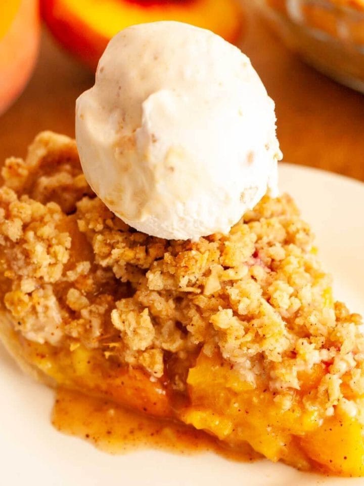 A slice of crumble top peach pie on a white plate topped with a ball of vanilla ice cream with peaches in the background.