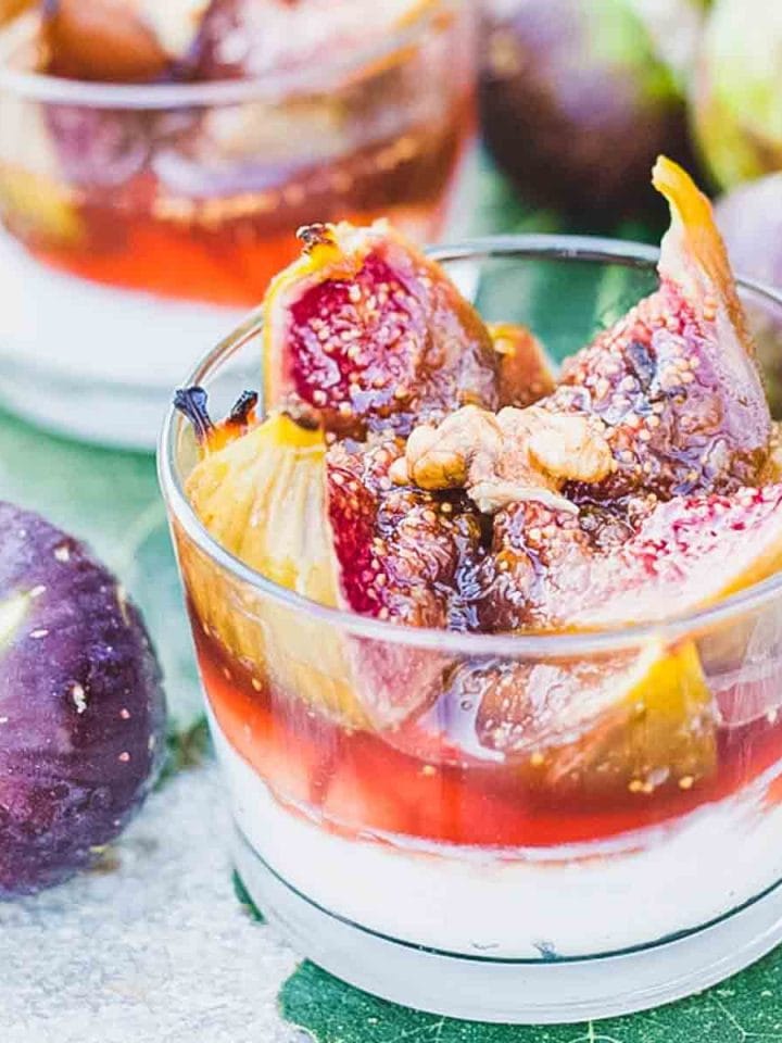 A close up of two glasses with honey roasted figs over Greek yogurt next to fresh figs and fig leaves.