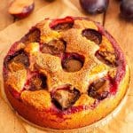 A round yogurt plum cake on a wooden table next to fresh plums.