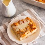A slice of bread pudding topped with vanilla sauce next to jug with vanilla sauce and baking dish.