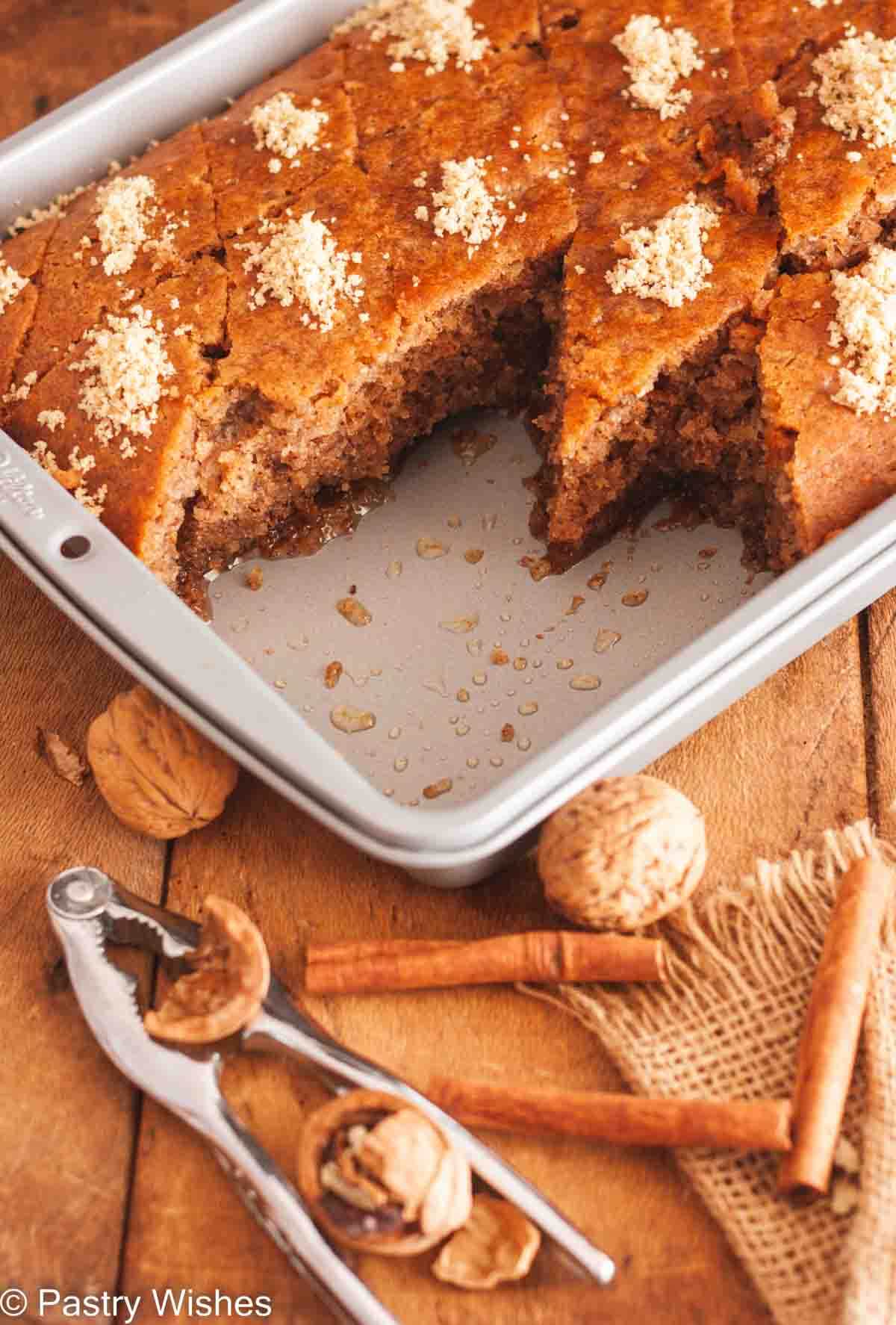A pan of diamond shaped karidopita on a wooden surface with walnuts, a nutcracker, and cinnamon sticks.