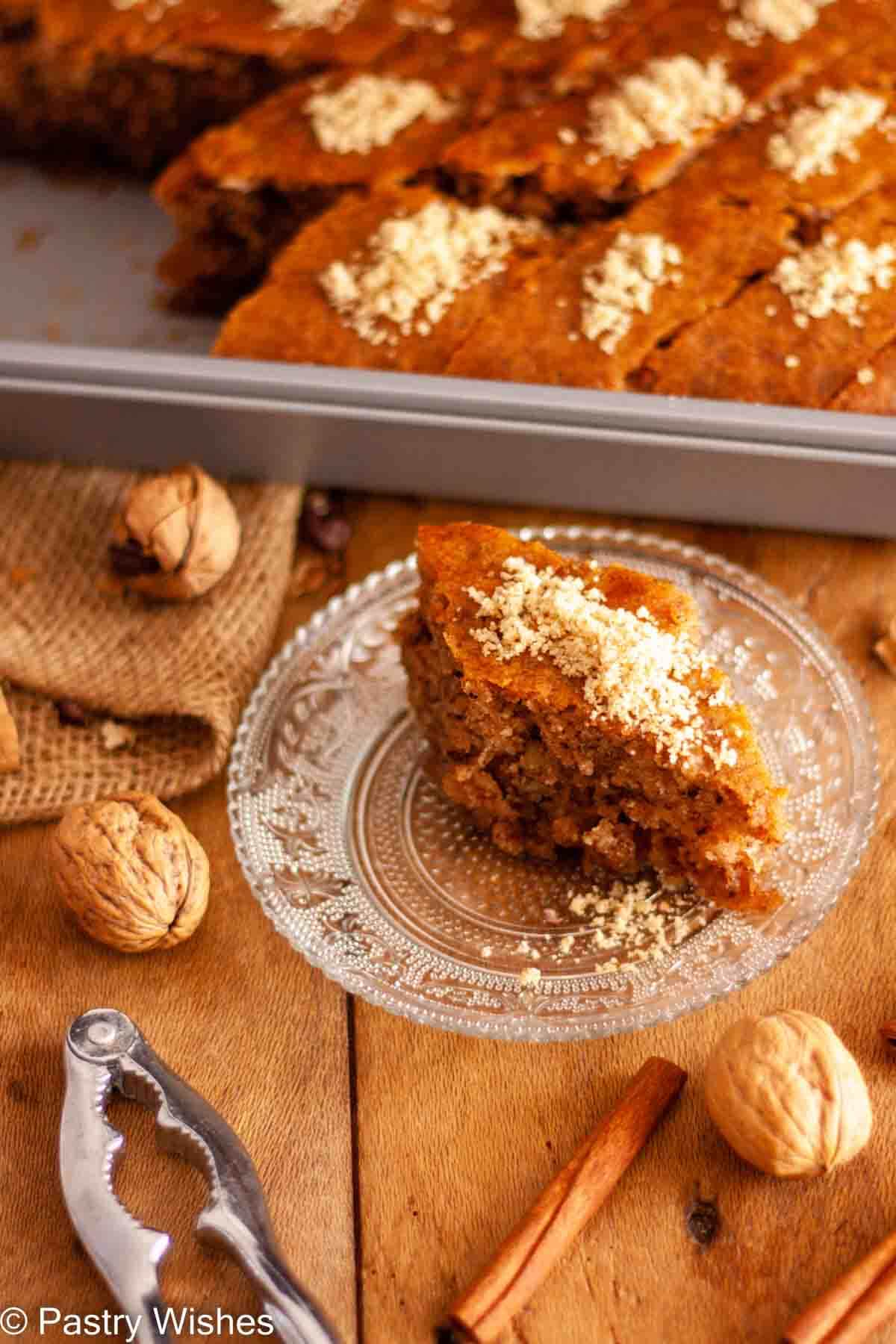 A slice of karidopita on a glass plate next to a pan of walnut cake on a wooden surface with walnuts, a nutcracker, and cinnamon sticks.