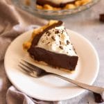 A slice of chocolate pie on a white plate with a fork in front of a pie plate with chocolate pie.
