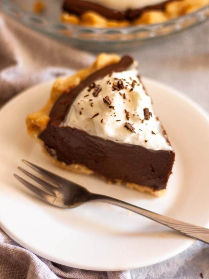 A slice of chocolate pie on a white plate with a fork in front of a pie plate with chocolate pie.