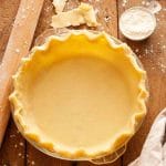 A closeup of an unbaked pie crust in a glass pie plate on a wooden table with flour, a kitchen towel, and a rolling pin.
