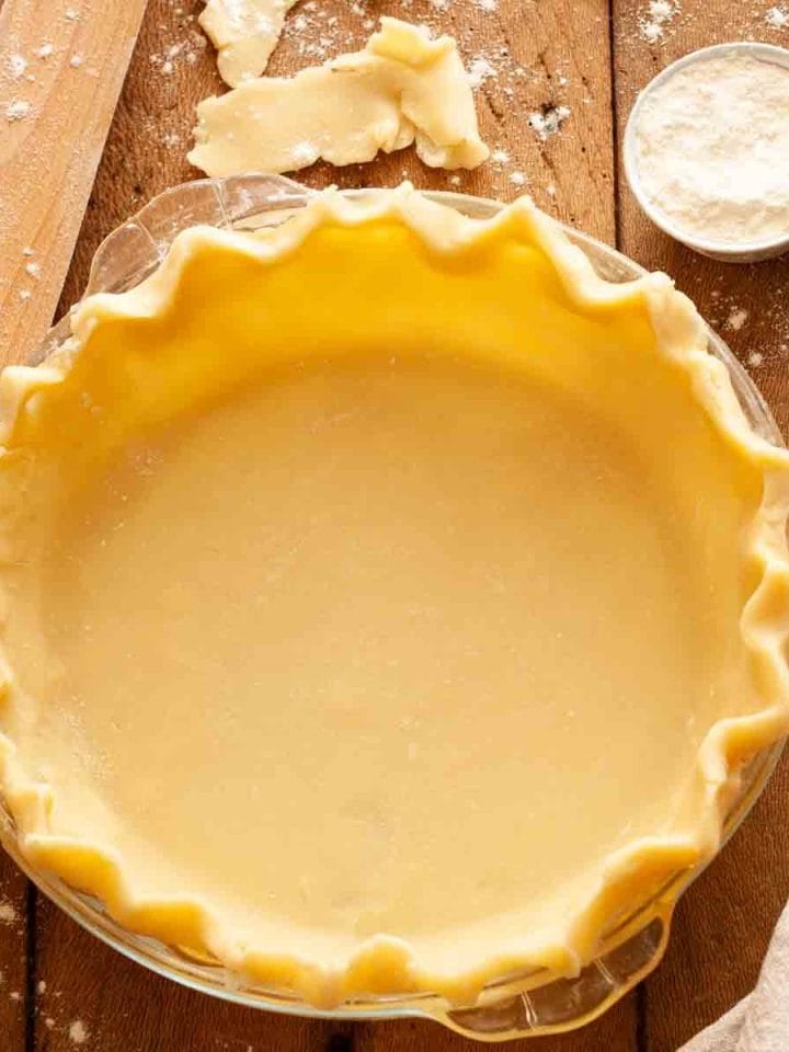 A closeup of an unbaked pie crust in a glass pie plate on a wooden table with flour, a kitchen towel, and a rolling pin.