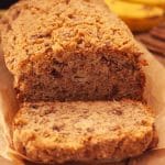 A close up of sliced banana bread with streusel topping with bananas and cinnamon sticks in the background.
