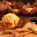 A close up of a cinnamon streusel muffin on burlap and a wooden table next to cinnamon sticks and tulip lined muffins.
