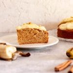 A slice of spiced banana cake on a white plate next to a banana and cinnamon sticks.
