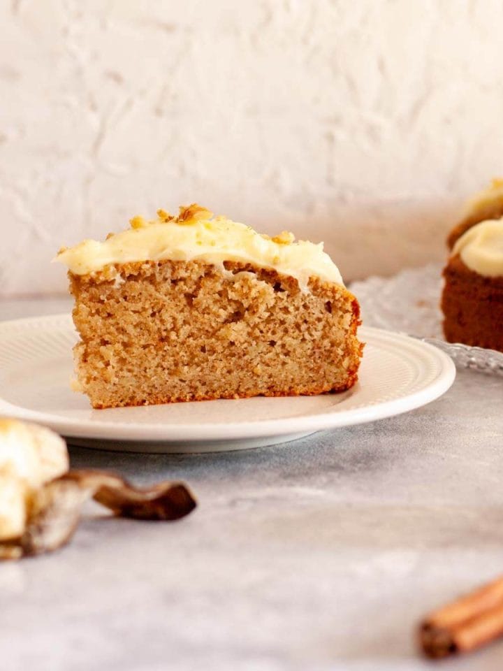 A slice of spiced banana cake on a white plate next to a banana and cinnamon sticks.