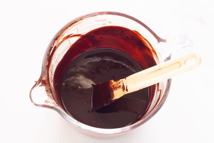A large glass measuring cup filled with a mixture of melted butter, vanilla, and cocoa powder and a small spatula.