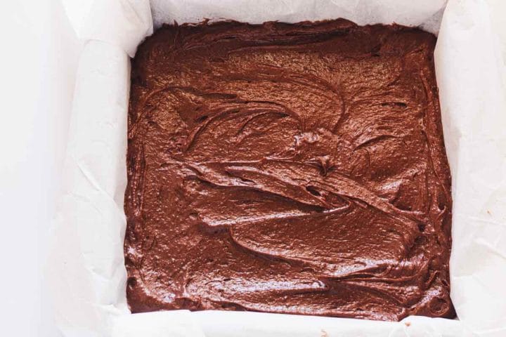 A square baking pan lined with overhanging parchment paper and filled with brownie batter.
