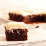 A close up of a brownie slice with cream cheese frosting on parchment paper with other brownies in the background.