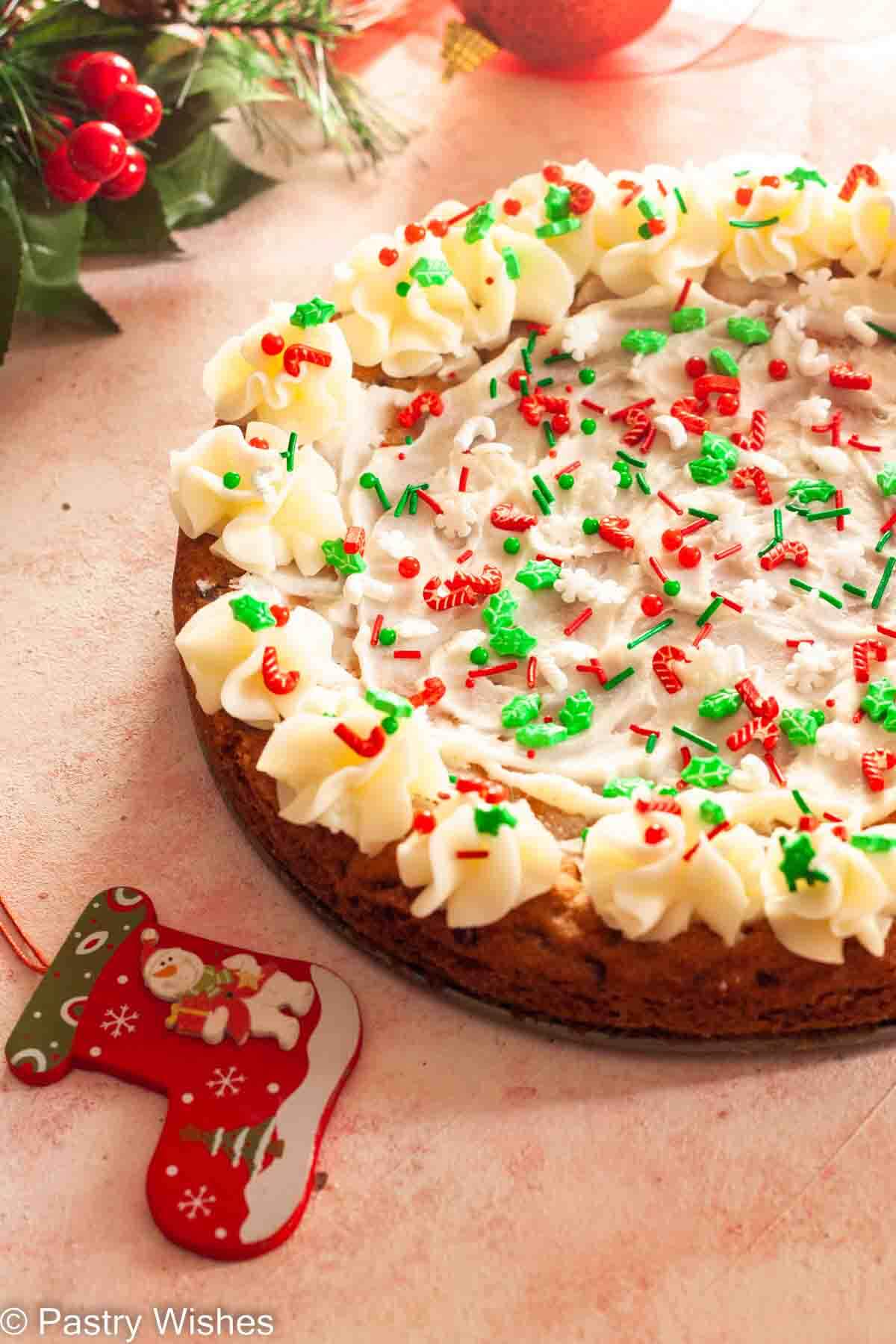 A Christmas cookie cake on a pink and white surface next to Christmas ornaments.