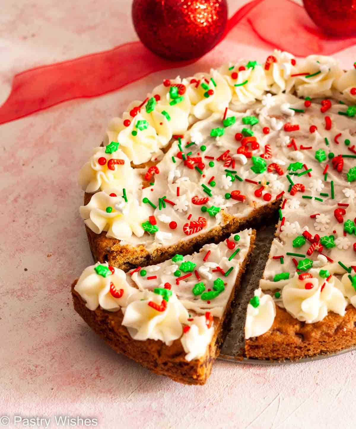 A sliced Christmas cookie cake on a pink and white surface with Christmas decorations in the background.