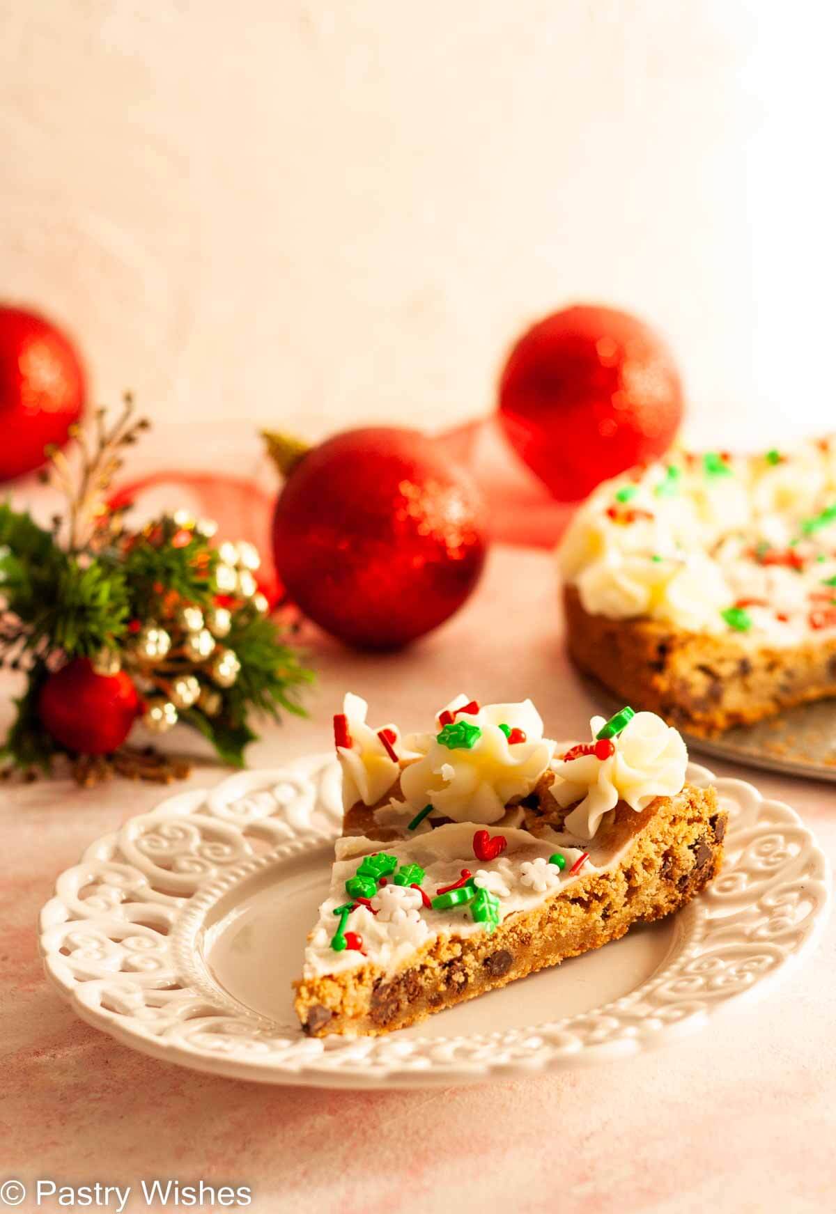 A slice of a Christmas cookie cake on a white plate with the remaining cookie cake and Christmas decorations in the background.