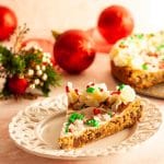 A slice of a Christmas cookie cake on a white plate with the remaining cookie cake and Christmas decorations in the background.