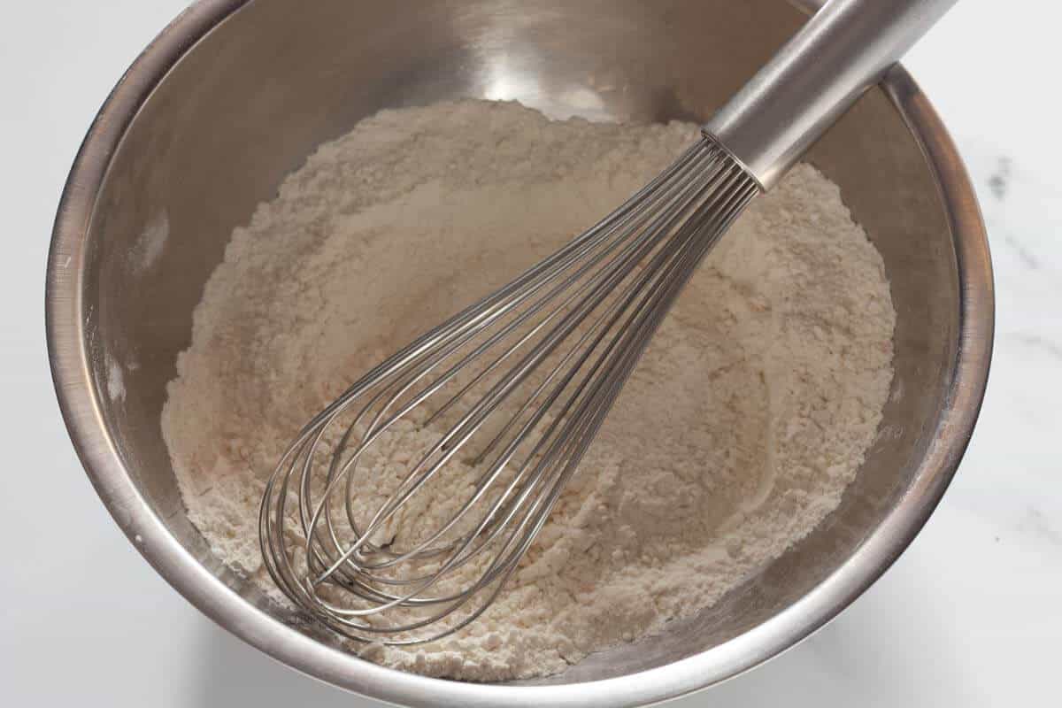 A metal bowl with flour, cornstarch, baking soda, salt and a whisk.