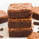 A stack of three thick brownie squares next to other brownies on a white surface.