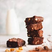 A closeup of a stack of dairy free brownies on a white surface next to chopped walnuts, chocolate, cocoa powder next to a brownie square with dairy free milk in the background.