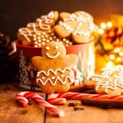 A close up of a gingerbread cookie on a candy cane on a wooden table with other cookies in a cookie box next to ornaments and lights.