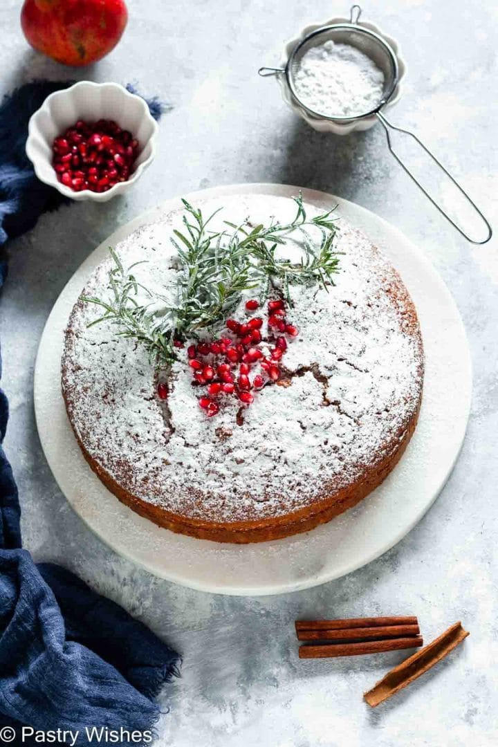 An overhead image of a greek new years cake topped with powdered sugar, rosemary sprigs and some pomegranate seeds, next to a bowl of pomegranate seeds, cinnamon sticks, and a mesh strainer with powdered sugar.