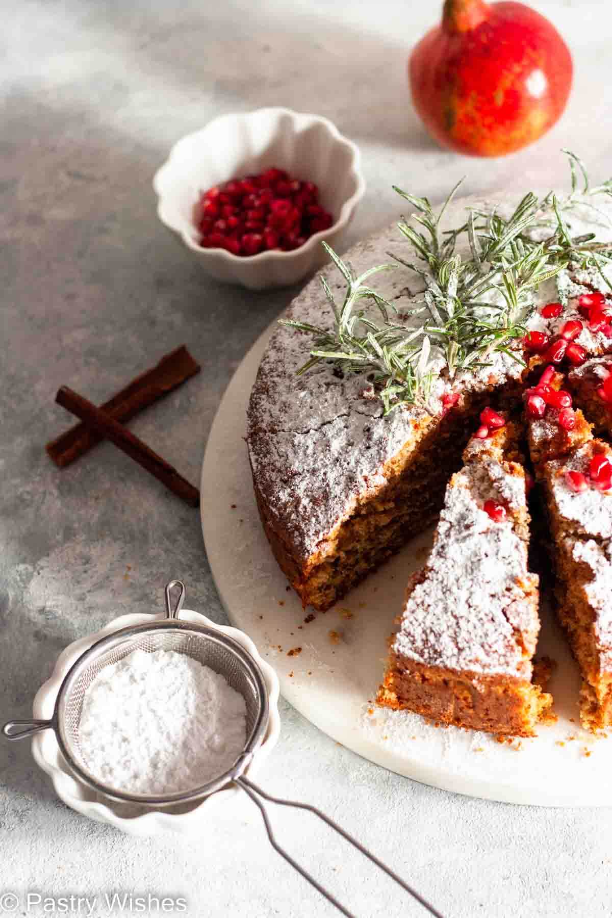 Sliced greek new years cake with two cut pieces on a white platter next to a bowl of pomegranate seeds and powdered sugar in a small mesh strainer.