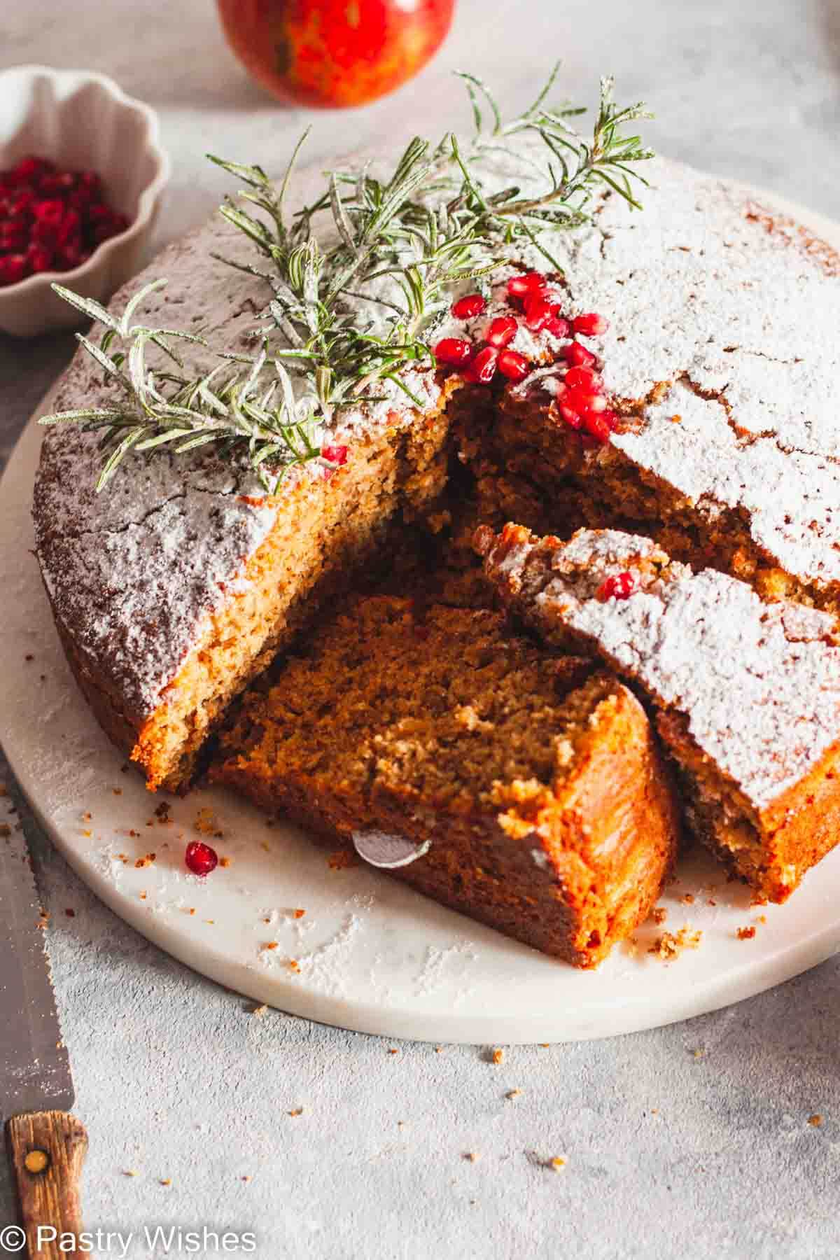 Sliced greek new years cake with two cut pieces on a white platter next to a bowl of pomegranate seeds and a knife.