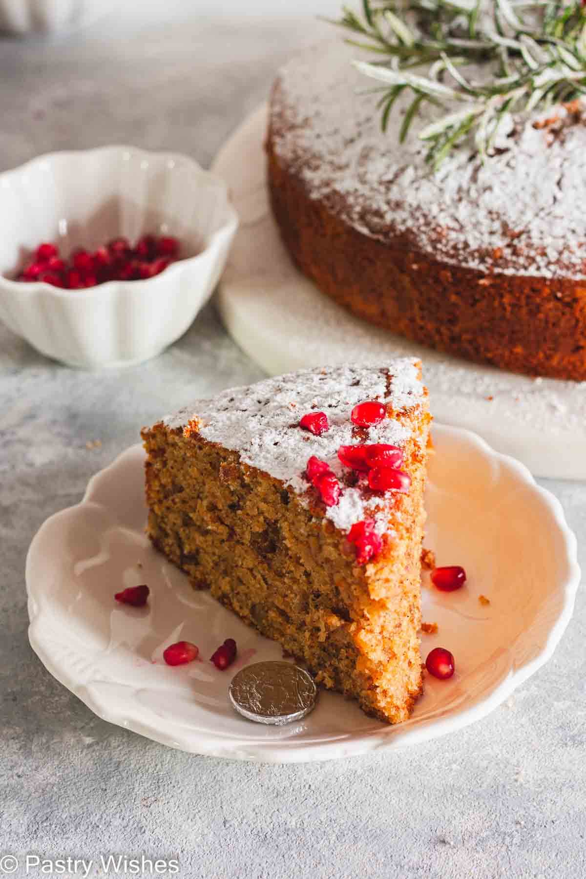 A slice of greek new years cake on a white plate next to pomegranate seeds and a coin wrapped in foil and cake in the background.