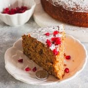 A closeup of a slice of greek new years cake on a white plate next to pomegranate seeds and a coin wrapped in foil and cake in the background.