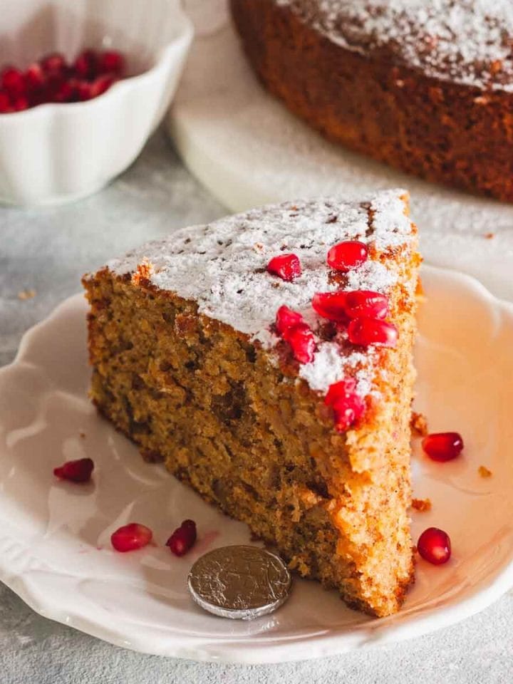 A closeup of a slice of greek new years cake on a white plate next to pomegranate seeds and a coin wrapped in foil and cake in the background.