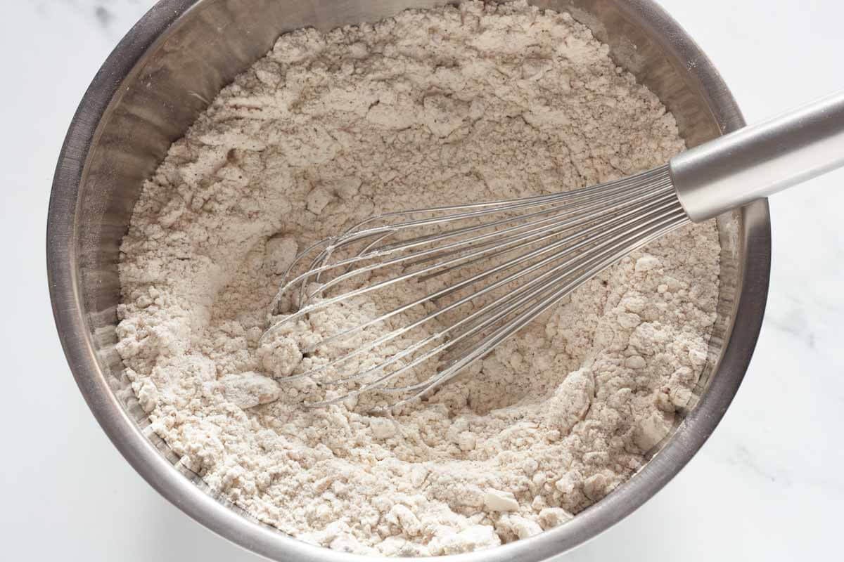 A mixing bowl with flour, spices, baking powder, walnuts and a whisk.