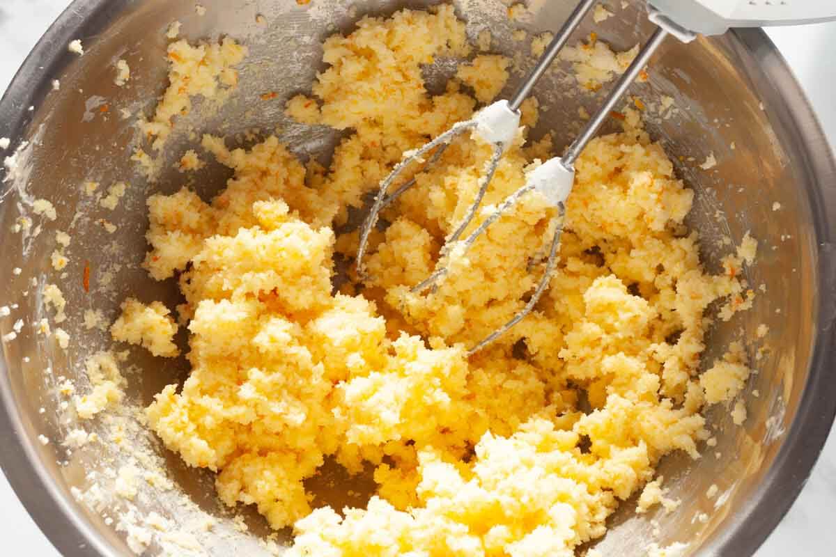 A hand mixer beating orange zest and sugar with butter in a metal mixing bowl.