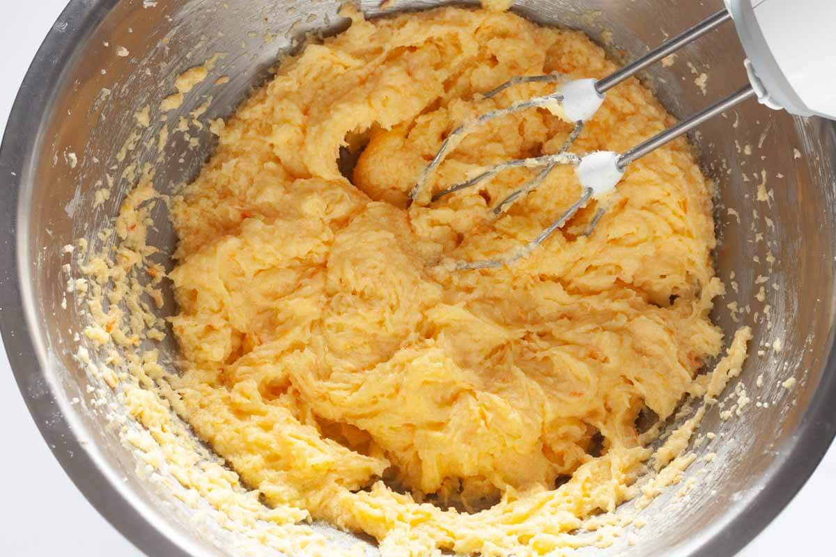 A hand mixer beating eggs, vanilla, orange zest, and sugar with butter in a metal mixing bowl.