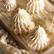 A closeup of meringue Christmas trees with edible gold glitter on a white and gold tray next to white beads and lace.