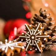 A close up of a snowflake gingerbread cookie next to a pine cone which Christmas lights in the background.