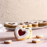 A closeup of a heart jam cookie in front of stacks of other cookies with a plate full of cookies in the background.