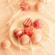 Pink and white Valentine cake pops on a white plate and pink and white surface next to a jar filled with sugar.