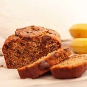 A closeup of sliced chocolate chunk banana bread on a white surface next to two bananas.