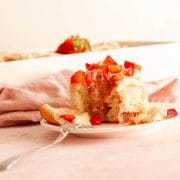 A close up of a strawberries and cream cinnamon roll opened with a fork and topped with chopped strawberries next to a pink kitchen towel and a baking dish.