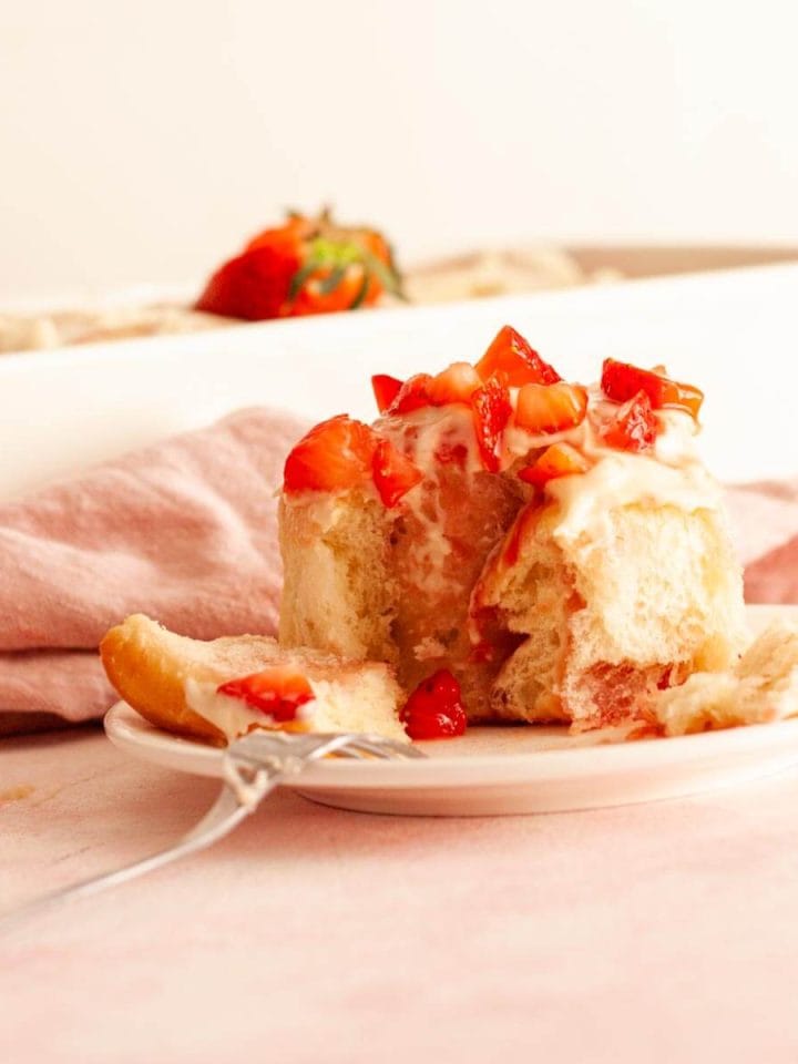 A close up of a strawberries and cream cinnamon roll opened with a fork and topped with chopped strawberries next to a pink kitchen towel and a baking dish.