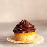 A close up of a vanilla cupcake with chocolate buttercream frosting and an paper liner pulled away.