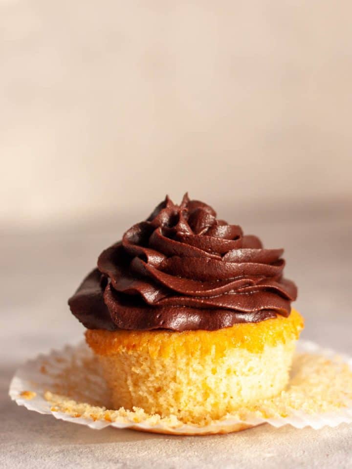 A close up of a vanilla cupcake with chocolate buttercream frosting and an paper liner pulled away.