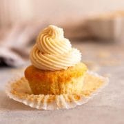 A closeup of a vanilla cupcake with an open paper liner topped with almond buttercream frosting.