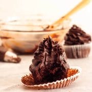 A close up of a bitten chocolate fudge cupcake with chocolate fudge frosting with a piping bag, a bowl and spatula in the background.