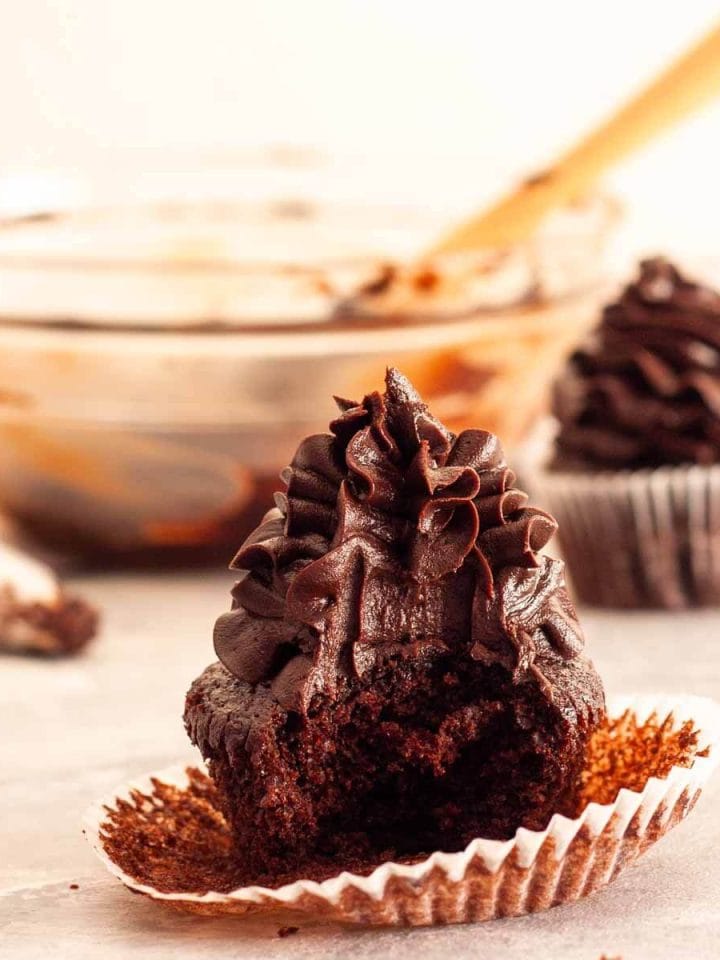 A close up of a bitten chocolate fudge cupcake with chocolate fudge frosting with a piping bag, a bowl and spatula in the background.